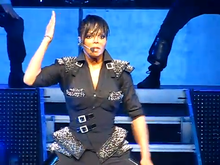 A woman on a stage, wearing dark clothing and a microphone; one of her arms is at her side while the other is extended into the air.
