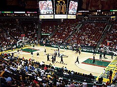 Interior of a basketball arena with a sparse crowd and empty red seats in many sections. A large video board hangs above the court, where players are warming up in small groups.