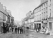 "Black and white photograph of the main street of the Irish town of Castlebar, with a group of people on the roadway to the left and pedestrians on the footpath to the right"
