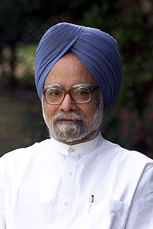 A portrait photograph of a bespectacled Indian man with a dark grey beard, a blue turban, and a white button-down shirt standing before a natural backdrop of trees. A pen is in his pocket.