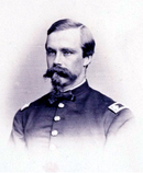 Black and white photo shows a man with neatly combed hair and a very bushy moustache and goatee. He wears a dark military uniform.