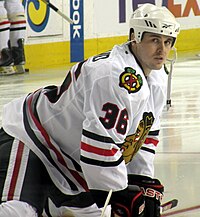 Hockey player in white uniform with a picture of crossed hockey sticks on the shoulder and the painted face of an indigenous person on the chest. He is crouched on the ice, feet further than shoulder width apart.