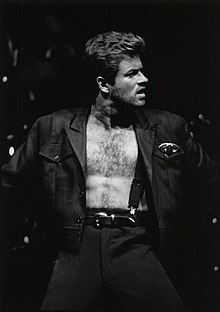 Michael dressed in a black leather suit, standing on a stage, holding out a microphone to the audience