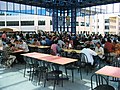 Grande et haute salle vitrée, à l’étage, tables de couleurs différentes selon les rangées et chaises[N 25].