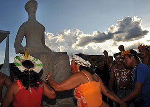 Indigenous people holding hands, two women in the foreground, surround a statue that represents Justice