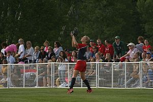 A light-skinned woman, wearing a red and blue shirt and red shorts, has her arms in the air to catch a ball in a grassy field as spectators look on.