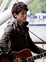 A three-quarter portrait of a male teen looking forward. He has dark, curly hair, wears a black leather jacket, and strums a guitar hung over his shoulder.