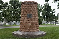 Photo d'un monument vertical composé de pierre rouge, avec une base plus large. Á l'arrière-plan sont visibles des tombes.