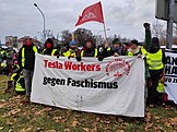 At an outdoor demonstration in Riesa, Germany, six Tesla workers carry a banner that reads "Tesla Workers gegen Faschismus" ("Tesla workers against fascism"). There is an IG Metall flag in background, and also on the banner itself. The faces of people holding the banner are blurred.