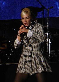 A color photograph of Gwen Stefani performing live against a purple-pink background.