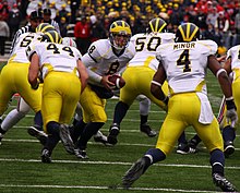 American football player in white, yellow and blue uniform prepares to hand the football to a teammate.