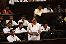 photograph of Wickramanayake dressed in a white saree surrounded by other people listening to her speech