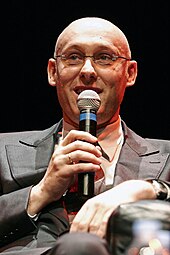 Photographie en gros plan d'un homme chauve, à lunettes, assis sur un fauteuil, parlant dans un micro et éclairé par un projecteur tirant sur le rouge.