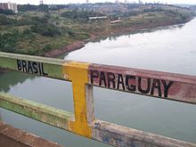 Photographie de la rambarde d'un pont en béton. Deux mots sont écrits en noir et en capitales : Brasil à gauche, sur un matériau peint en vert, Paraguay à droite, sur un matériau peint en rouge. Une barre verticale jaune sépare les deux mots. Derrière la rambarde, on peut voir un fleuve, et en arrière plan des champs et des immeubles sur une des rives du fleuve.