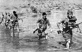 a black and white photograph of a line of males in uniform carrying weapons wading through a shallow river