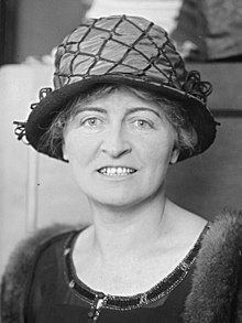 White woman, seated, smiling, in 1922. She is wearing a dark dress, a fur stole, and a hat.