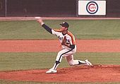 A man in a white baseball jersey with "ASTROS" on the chest and orange and yellow stripes on his shoulders pitches a baseball with his right hand.