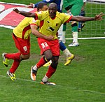 Two men wearing yellow shirts, red shorts and red socks, celebrating on a grass field