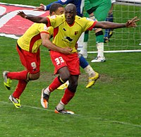 Two men wearing yellow shirts, red shorts and red socks, standing on a grass field. Both appear to be celebrating: one man has his arms aloft, the other is following him.