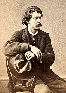 Black and white photograph of a moustachioed man, crouching, wearing a large hat in his left hand.