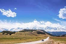 Photographie lumineuse montrant une voiture sur la route, avec en arrière-plan des sommets enneigés du massif de la Tchouïa du Nord. La route évite une colline qui se situe au centre, et se dirige vers la droite de l'image.