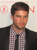 A man dark hair wearing, black suit, including black tie and a plad shirt.