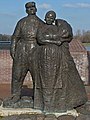 Photographie en couleurs d'une sculpture montée sur un socle en pierre et d'une voie d'eau en arrière-plan