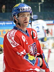 An ice hockey player standing, looking to the right of the camera. He is wearing a blue helmet and a red uniform.