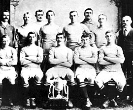 A group of thirteen men, eleven in association football attire typical of the early twentieth century, and two in suits. A trophy sits in front of them.