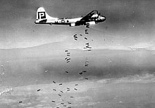 Black and white photograph of a World War II-era bomber releasing bombs. The bombs are falling in a scattered pattern.