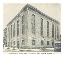 This is a black-and-white photo of the Ludlow Street Jail, a cuboid brick building visible here on two sides. Ten arched windows, five large and five small, are each on both sides. A sidewalk wraps around the building. The photo is originally captioned in all-caps, "Ludlow Street Jail, Ludlow and Exeter Streets".