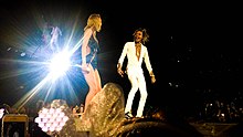 Two people—a young white woman in a sparkling black dress and heels, and a black man in top-to-toe white suits and sneakers, performing