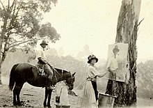 Photographie en noir et blanc. Au premier plan, l'artiste regarde le photographe tout en ayant à la main un pinceau à hauteur de la toile qui est suspendue à un arbre. Au deuxième plan, un homme à cheval de profil. En arrière plan, le bush australine.