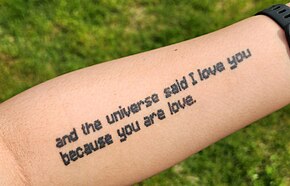 A pale white arm, tattooed with the quote "The universe loves you because you are love", all lowercase and in a Minecraft-like font.