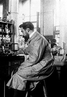 Photographie d’un homme brun, portant barbe et moustache, vu du côté gauche, assis sur un tabouret haut, devant une paillasse de laboratoire.