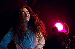 A half-waist picture of Lorde performing in a white dress shirt with a drummer and pink spotlight in the background.