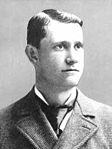 A photograph of a man who looks to the right of the image. He is shown from the upper chest to the top of his head. His hair is parted on the right side of his head, and he is wearing a suit and a tie with a large knot.