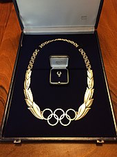 Metallic chain including the five Olympic rings, flanked on either side by olive wreaths, and a lapel badge including a smaller version of the five Olympic rings and olive wreaths