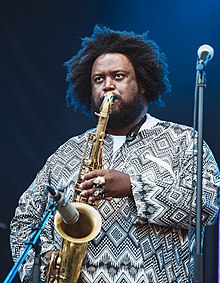A big man with finger bling wears an artistic black-and-white African-style shirt and puffs his cheeks blowing into a tenor sax, captured by a standing mic.