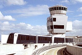 A four-car train traveling on top of a building with an empty concrete guideway in the foreground. An airport control tower is in the background, in front of a blue, cloudy sky.