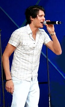 Basshunter, who is dressed in white, sings into microphone while standing in front of people and blue background