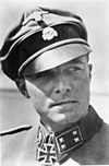 A man wearing a military uniform, peaked cap and a neck order in the shape of a cross. His cap has an emblem in shape of a human skull and crossed bones.