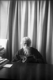 An old bearded man garbed in a dark mantle is reading from a slim book perched in his hands. He is sitting at a dark-toned desk cleared of everything but a neat stack of papers at left; in the background is a light-coloured curtain.