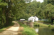 Photograph of a dirt towpath beside a canal with a lock with a house in the background