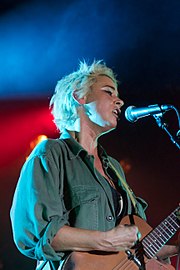 A blonde, short-haired woman wearing a green shirt performs with a microphone and guitar