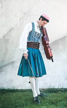 A tall, slender, white person standing in grass, wearing Bäckadräkten, and holding a nyckelharpa