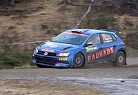 Five-door two-box car in a racing livery fitted with a rear aerofoil and a roof-mounted air scoop for ventilation travelling at speed on a gravel course