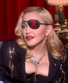 Madonna, wearing a black eyepatch with a jeweled X and dressed in a black hooded robe with jeweled crosses, sings to a microphone.
