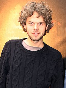 Three-quarter body shot of a 33-year-old man. He stands facing the camera with his right shoulder raised and hands placed into his jeans' pockets. He wears a dark jumper over a pale tee-shirt. His brown hair is slightly curly and about collar length. He has a slight beard.