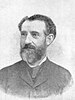 Head and shoulders of a dark-haired, bearded man of about 50 wearing a dark jacket and tied topped by a white collar.